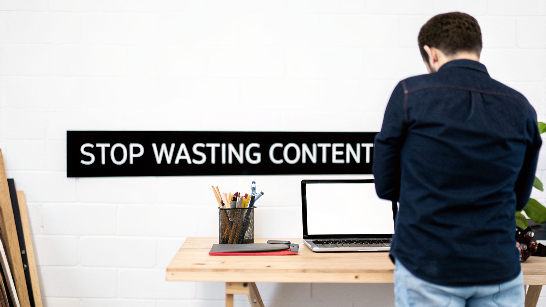 A man stands at a wooden desk with a laptop, facing a sign reading "STOP WASTING CONTENT" on a white brick wall.